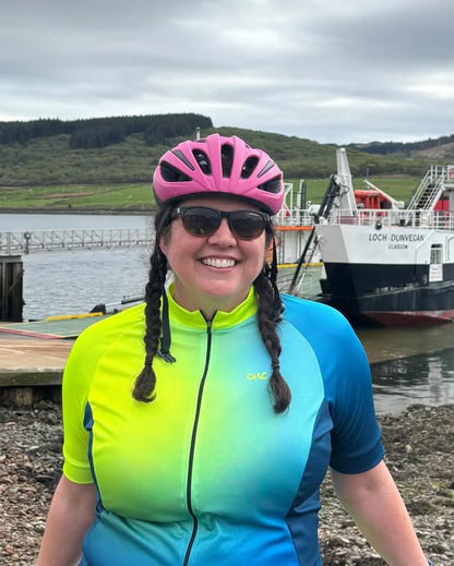Person in cycling gear with a pink helmet and sunglasses, standing on a pebbly shore with a ferry in the background.