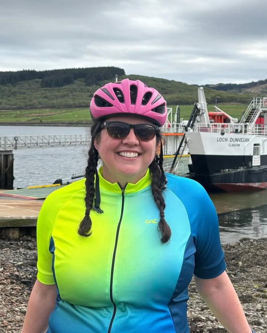 Person in cycling gear with a pink helmet and sunglasses, standing on a pebbly shore with a ferry in the background.
